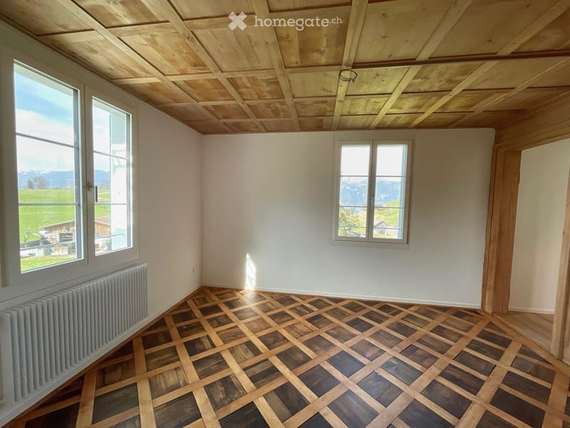 Empty room with wooden parquet flooring, white walls, exposed wooden ceiling beams, and large windows offering a scenic mountain view.