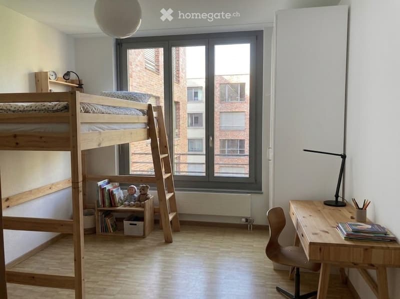 Bright, modern bedroom with a wooden bunk bed, desk, chair, and small shelf holding books and toys. Large window offers a view of the building outside.