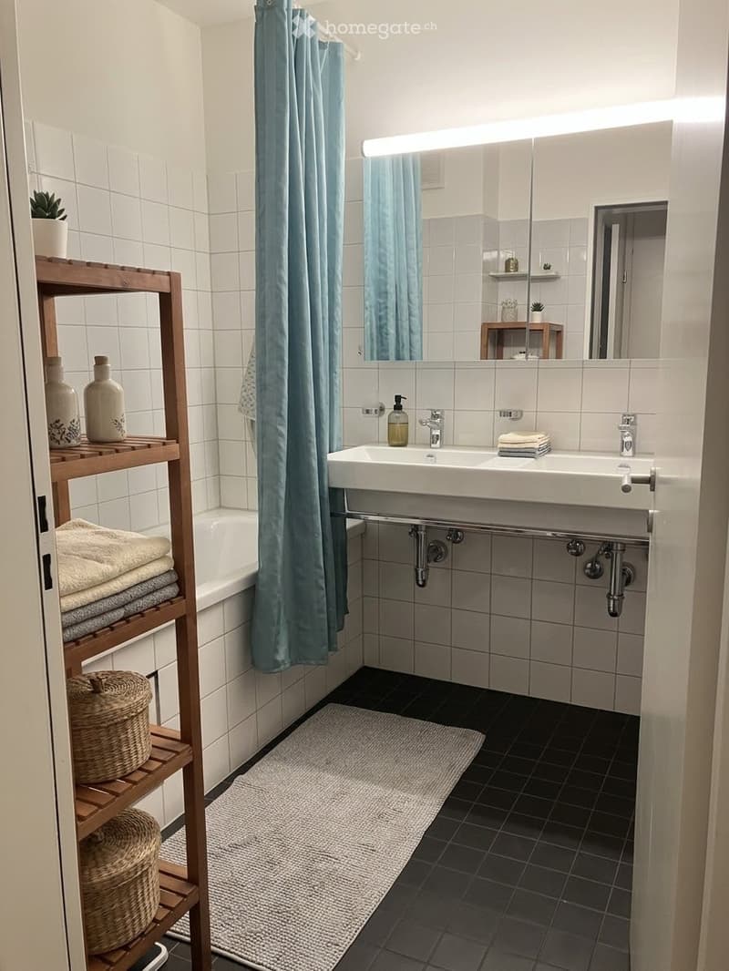 Modern bathroom with white tiles, double sink, bathtub, wooden shelf with towels and decorative items, and a blue shower curtain.