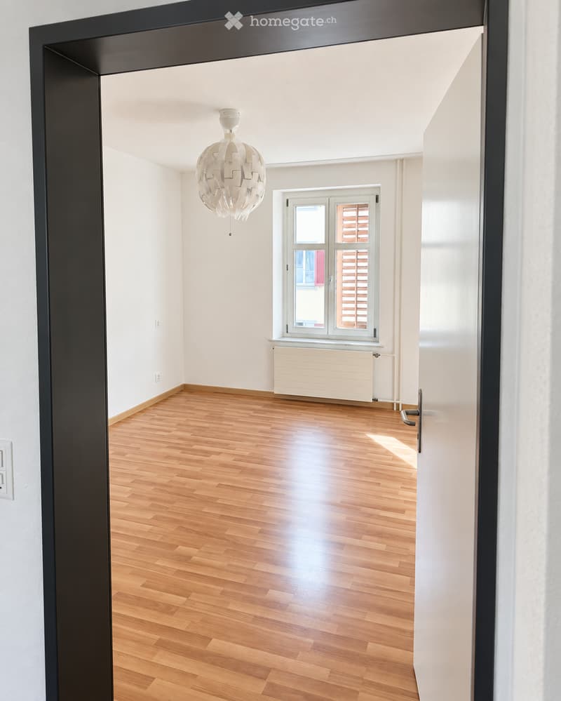 Empty room with wooden flooring, white walls, a window with blinds, a radiator, and a modern pendant light fixture.