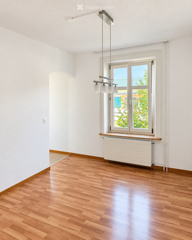 Empty room with wooden flooring, white walls, large window with view of greenery, radiator, and modern pendant light fixture.