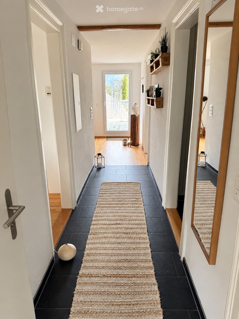 Modern hallway with dark tiled flooring, white walls, wooden trim, and a long woven runner. Features include a large mirror, small decorative items, and a view to an outdoor area through a glass door.
