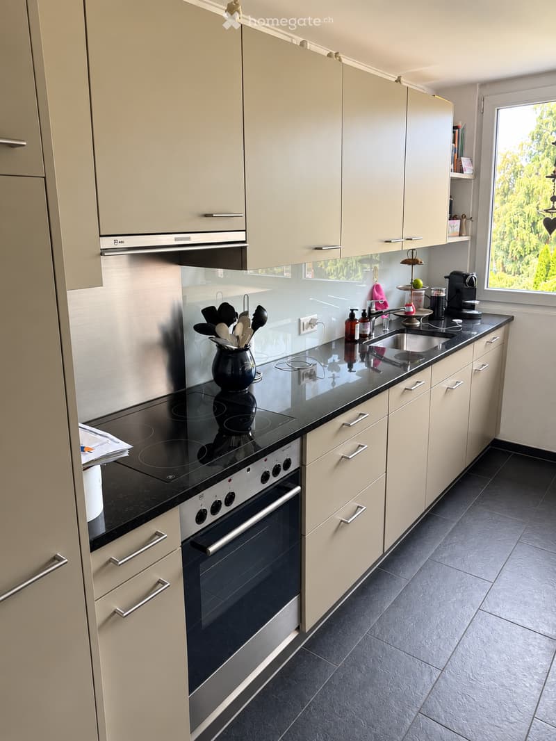 Modern kitchen with light-colored cabinets, black countertops, stainless steel appliances, and a large window offering a view of greenery.