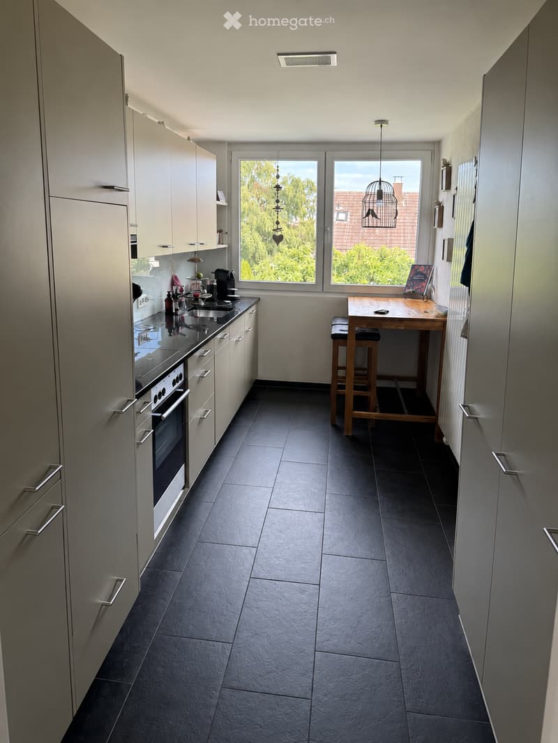 Modern kitchen with grey cabinets, dark tiled floor, and a central island with a table and stools. Appliances include an oven and coffee maker, with a window offering a view of greenery.
