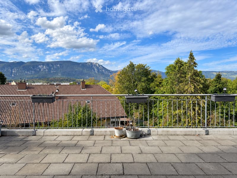 Balcony with metal railing, tiled floor, and potted plants, overlooking a cityscape with red-tiled roofs and distant mountains.