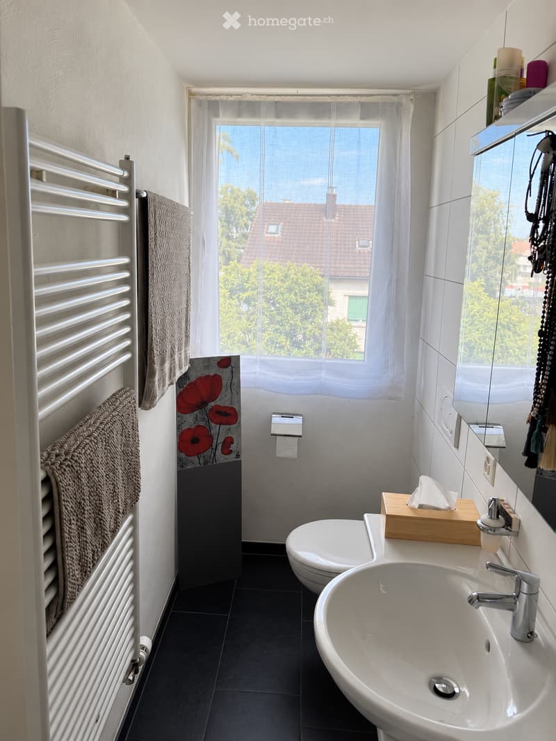 Modern bathroom with white tiles, a large window, a sink with a modern faucet, a toilet, a radiator with towels, a mirror, and a decorative wall with a floral design.