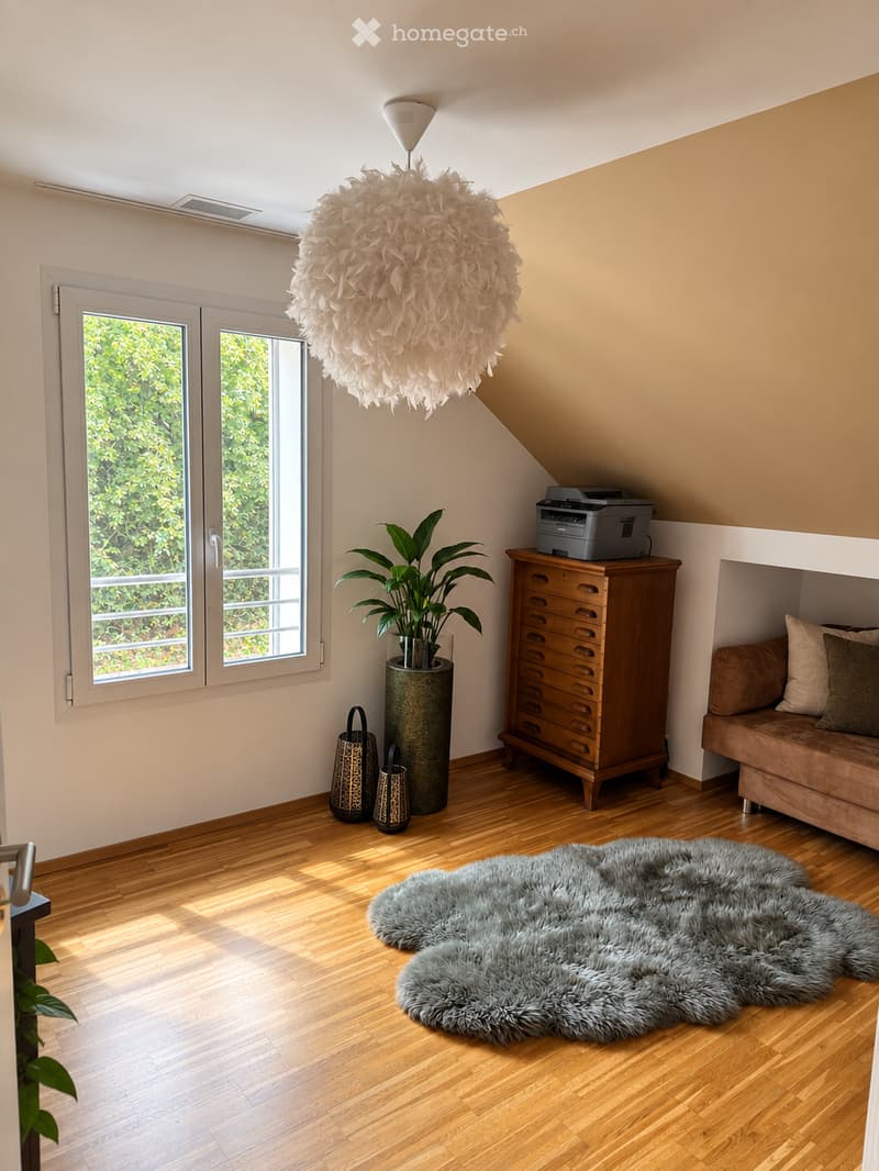 A cozy living room with wooden flooring, a large fluffy white hanging light fixture, a brown sofa with pillows, a wooden cabinet with drawers, a printer on top, a large furry rug, a tall potted plant, and decorative vases near the window.
