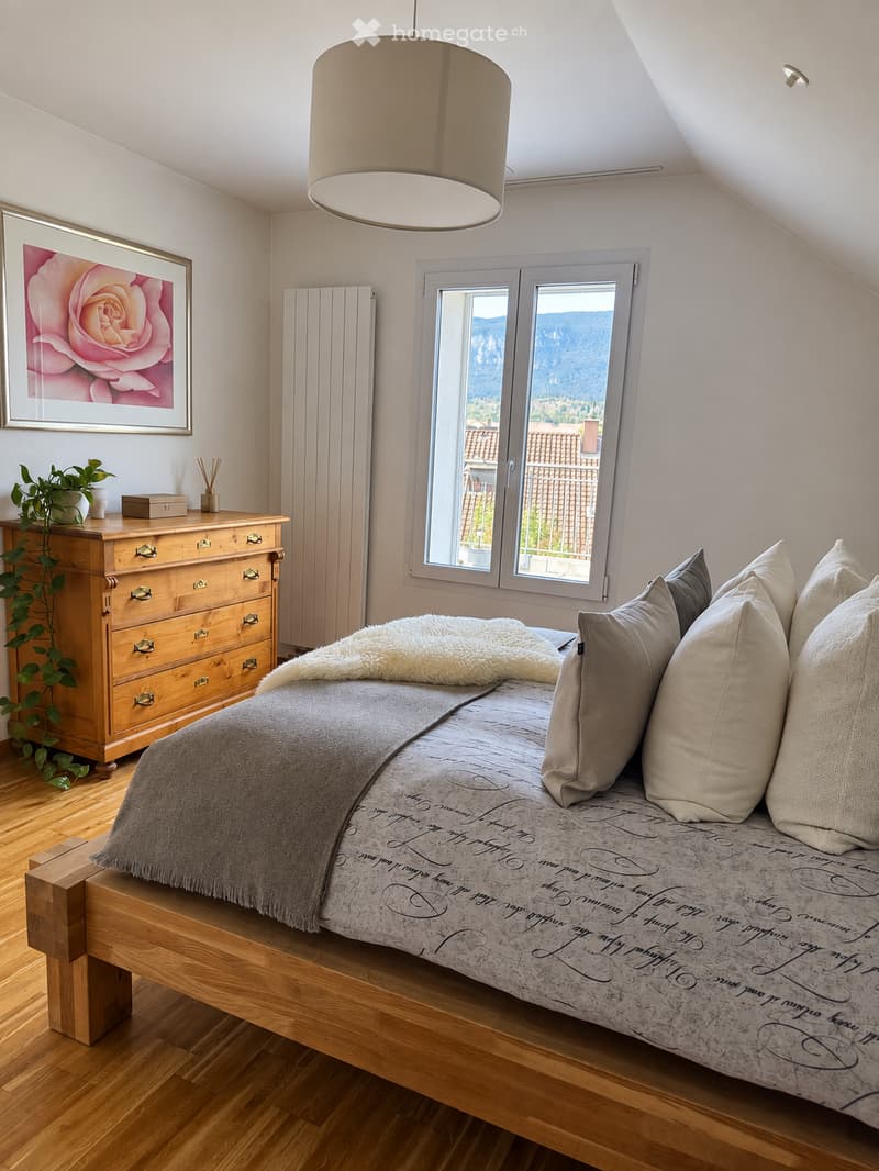 Cozy attic bedroom with wooden flooring, large window offering a scenic view, a wooden bed with a decorative bedspread and pillows, a wooden dresser with a plant, and a framed picture on the wall.