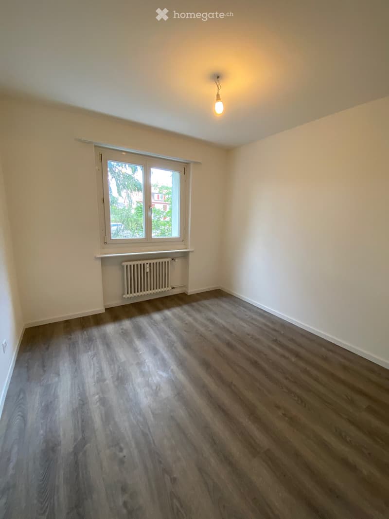 Empty room with wooden flooring, white walls, a window with a radiator below, and a single hanging light fixture.