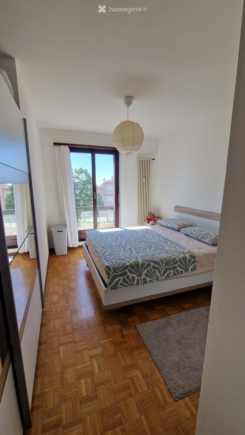 Bright bedroom with wooden flooring, large window with white curtains, bed with patterned duvet, nightstand with a toy, and a hanging paper lamp.