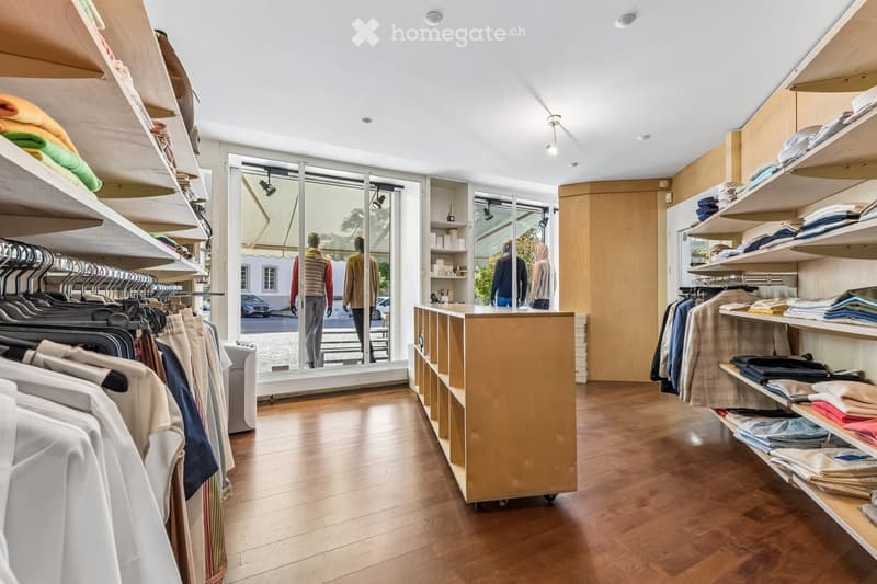 A well-lit clothing store with wooden flooring, clothing racks, shelves filled with folded garments, and mannequins near the entrance.