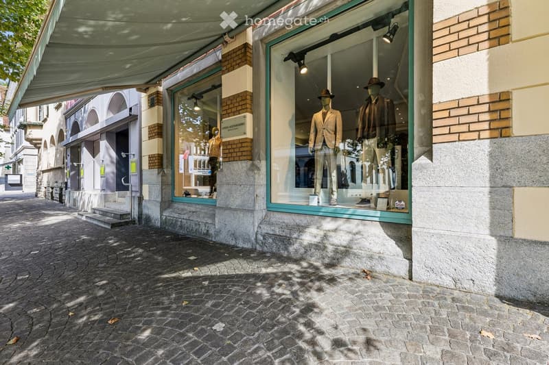 Facade of a building with large glass windows displaying mannequins in suits and hats, with a cobblestone street in front.