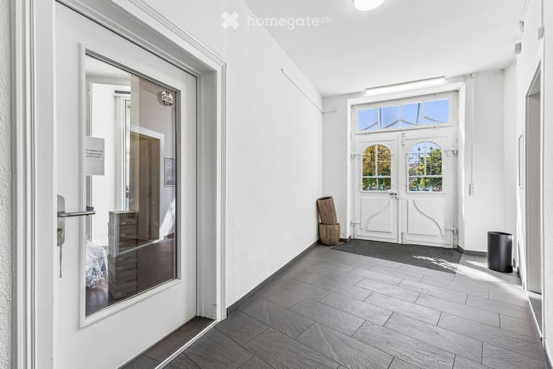 Bright hallway with white walls, tiled floor, and a large window at the end. Features a glass door with a view into another room, a small wooden vase, and a black trash bin.