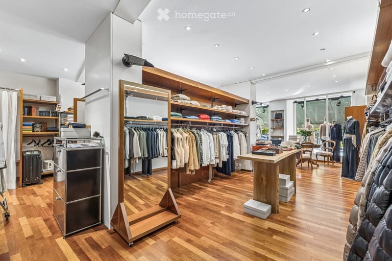 A well-lit clothing store with wooden flooring, clothing racks, shelves filled with folded garments, a wooden table with items on it, chairs, and a mirror. The space is organized and modern with large windows allowing natural light.