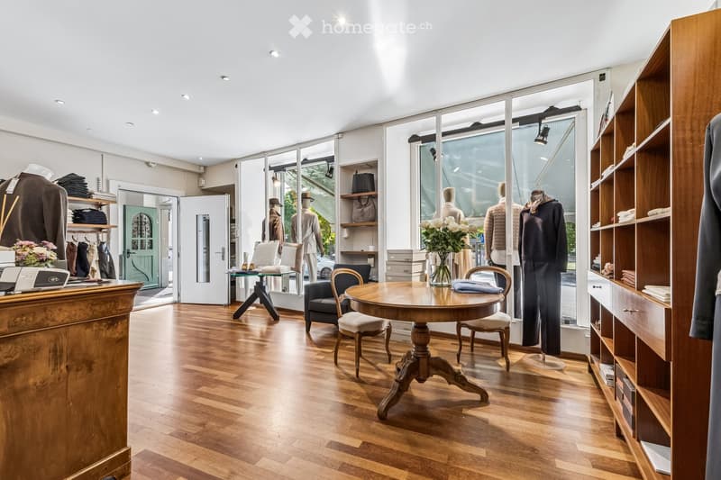 A well-lit, modern retail space with wooden flooring, large glass windows, and a round wooden table with chairs. Mannequins display clothing, shelves hold folded garments, and a cash register sits on a wooden counter.