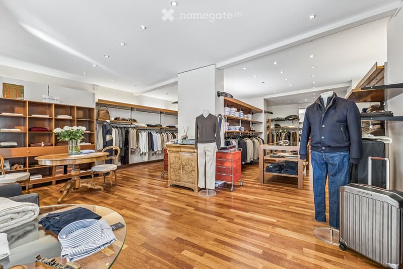 A well-lit clothing store with wooden flooring, shelves filled with folded clothes, a clothing rack, mannequins displaying outfits, a table with folded clothes, a rolling cart, and a suitcase near the entrance.
