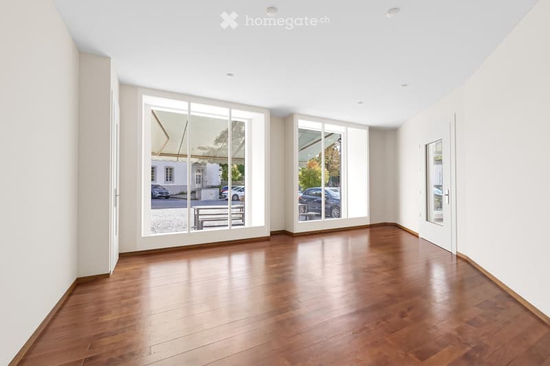 Empty room with wooden flooring, white walls, large glass doors, and a view of the street and parked cars.