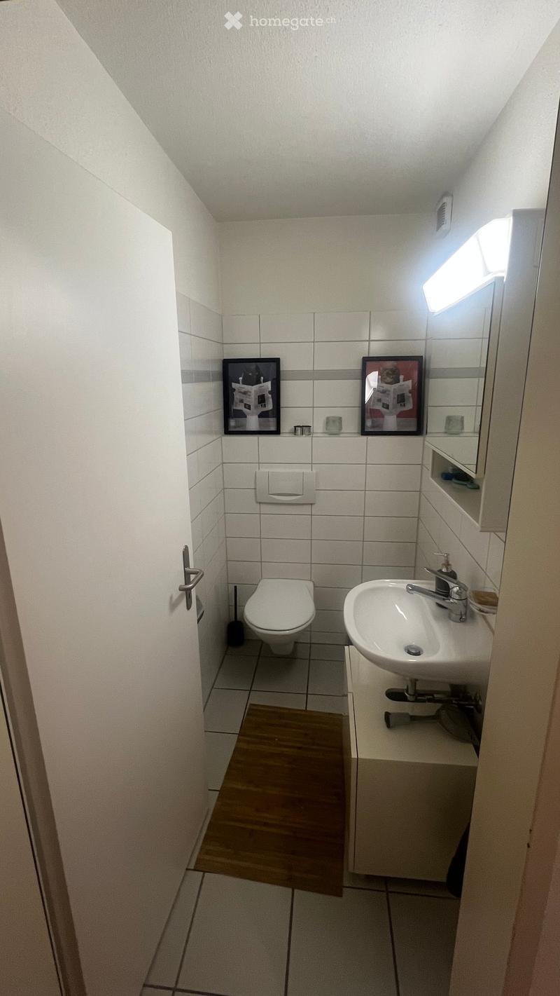 Compact bathroom with white tiled walls, a toilet, a sink with a mirror cabinet, and a small wooden floor mat.