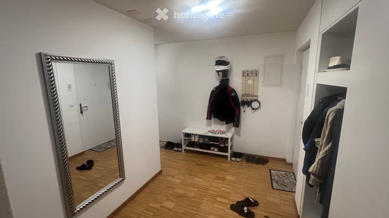 The image shows a well-lit hallway with wooden flooring, white walls, and a large mirror on the left side. There is a coat rack with jackets and a helmet, a small shoe rack with several pairs of shoes, and a closet with hanging clothes on the right side.