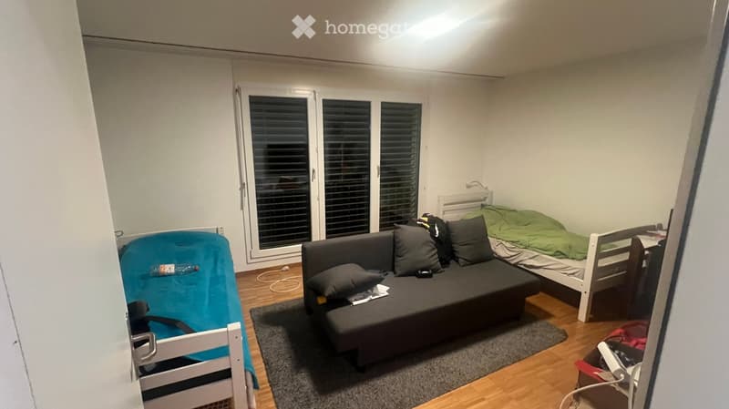 A small, furnished room with a sofa, a single bed, a chair, a small table, and a rug. The room features large windows with blinds and wooden flooring.