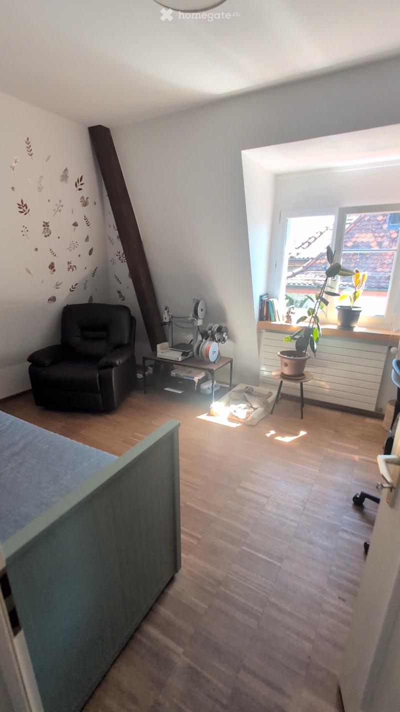 Cozy attic room with wooden flooring, slanted ceiling, and large window. Features a black leather sofa, small table with headphones and laptop, potted plants, and radiator.