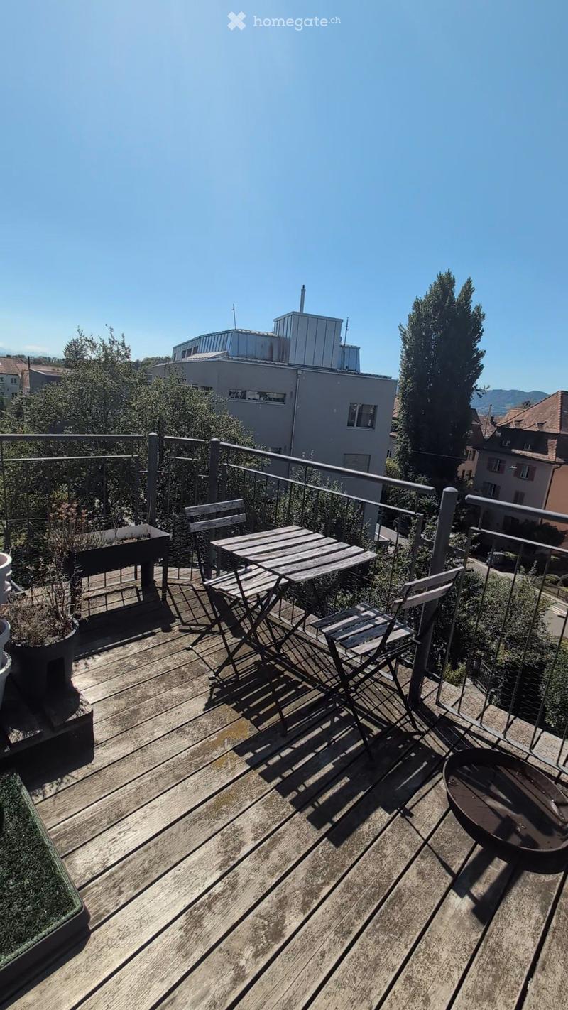 Wooden deck with a small table, chairs, potted plants, and a fire pit, overlooking a cityscape with buildings and trees.
