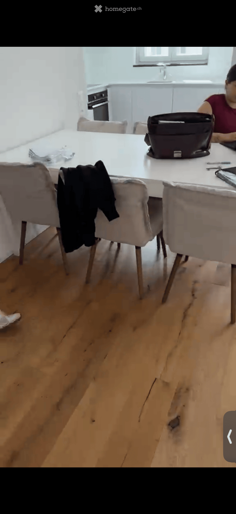 Dining area with a white table, four chairs, a laptop, and a backpack on the table. Wooden flooring and white walls.