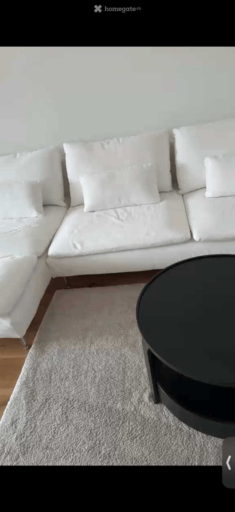 A modern living room with a white sectional sofa, multiple white cushions, a grey shag rug, and a black round coffee table.