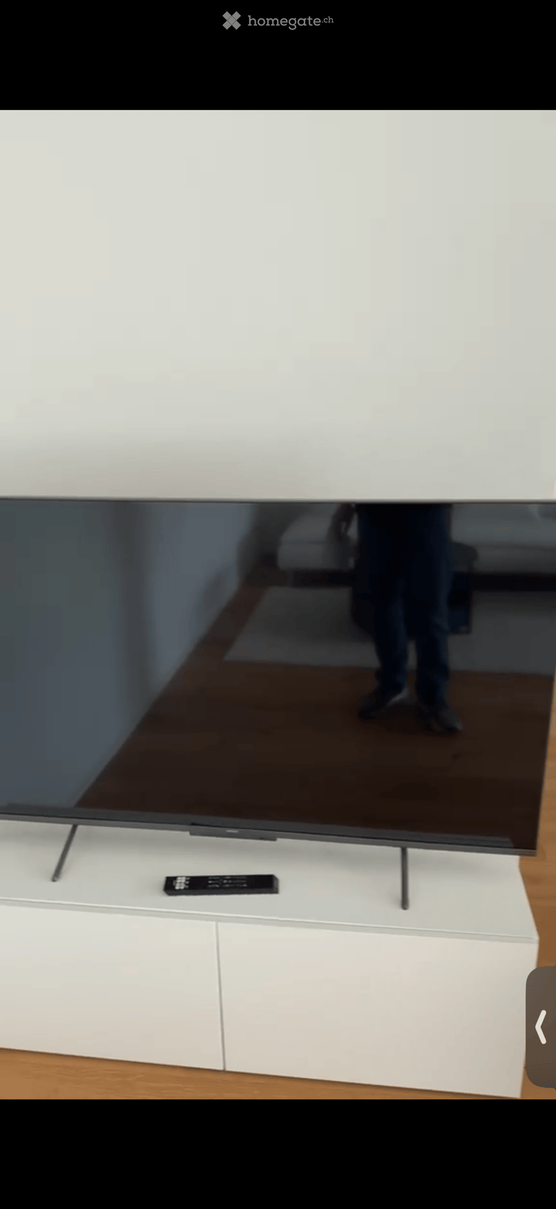 A modern living room with a large flat-screen television on a white TV stand, accompanied by a remote control. The room features wooden flooring and white walls.