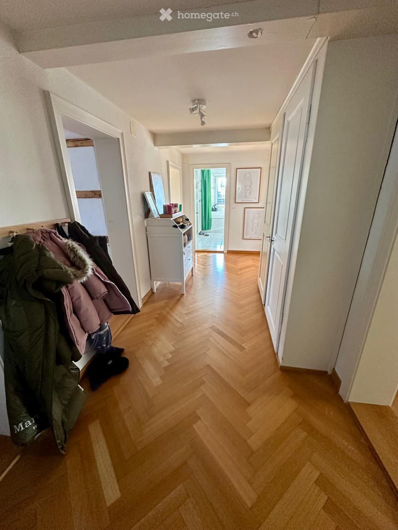 A well-lit hallway with wooden herringbone flooring, white walls, and several doors leading to different rooms. A small white cabinet with drawers and a mirror is visible, along with coats hanging on a rack.