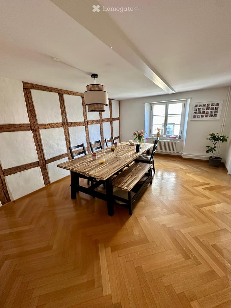 Bright dining room with wooden floor, large wooden dining table, bench seating, chairs, vase with flowers, and a potted plant near the window.