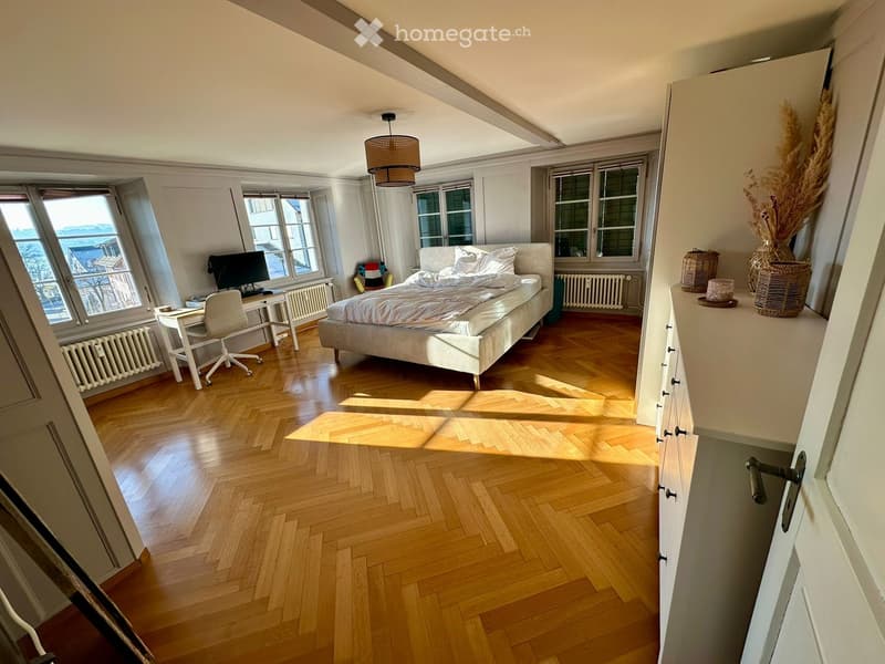 Bright bedroom with wooden herringbone flooring, large windows, a bed with white bedding, a desk with a chair, and a screen. A vase with dried plants sits on a white cabinet.