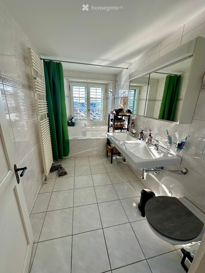 Bathroom with white tiles, double sink, mirror, bathtub, radiator, and a small stool. Green curtains cover the window and shower area.