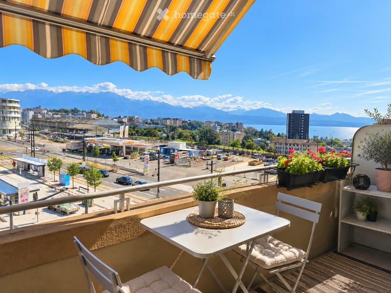 Balcony with a table, chairs, and potted plants, overlooking a cityscape with mountains in the background.
