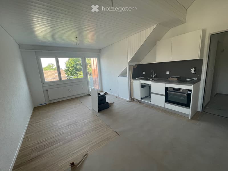 Empty kitchen with white cabinets, a sink, and an oven, featuring wooden flooring and large windows with a view of greenery outside.