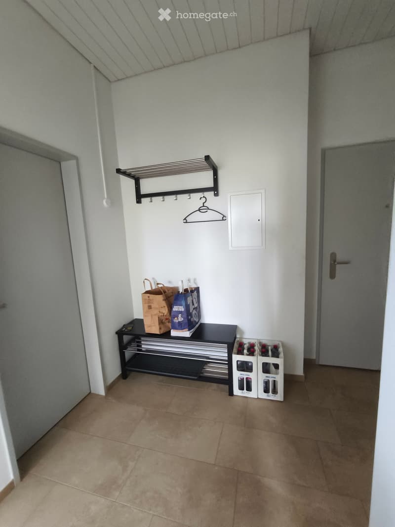 A small hallway with a tiled floor, a black metal table with two bags on it, a bottle rack with several bottles, and a coat rack with a single hanger.