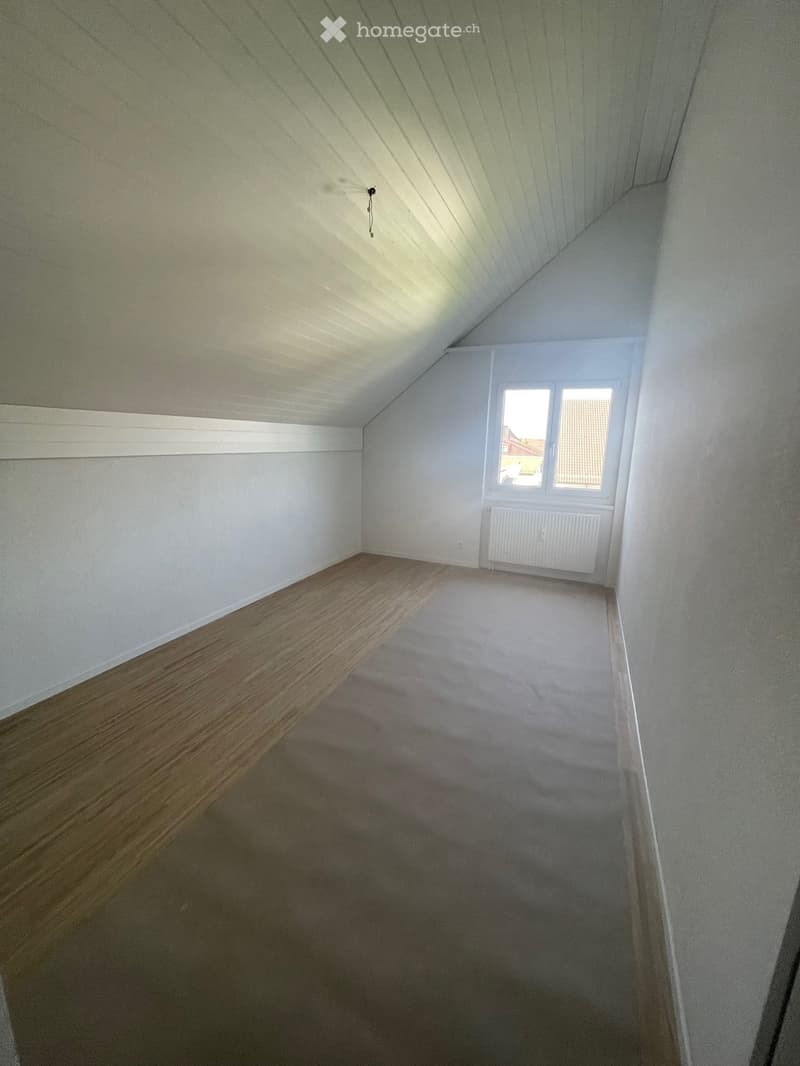 Empty attic space with wooden flooring on one side and carpet on the other, white walls, and a slanted ceiling with exposed beams. A window with white frame and a radiator below it are visible.