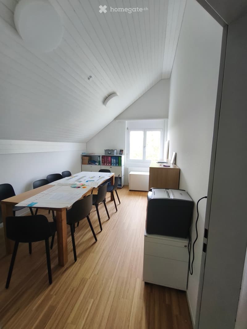 Bright attic room with wooden flooring, large window, table with chairs, printer, and shelves.
