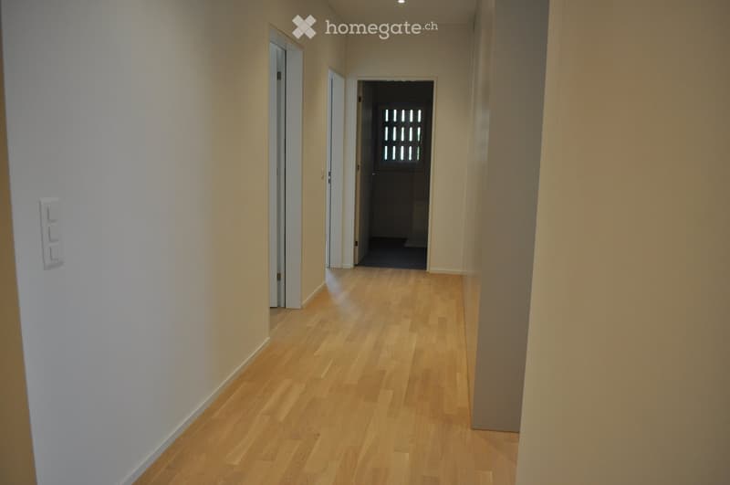 A clean, well-lit hallway with light wooden flooring and white walls. The corridor features several doors and a switch panel on the left side.
