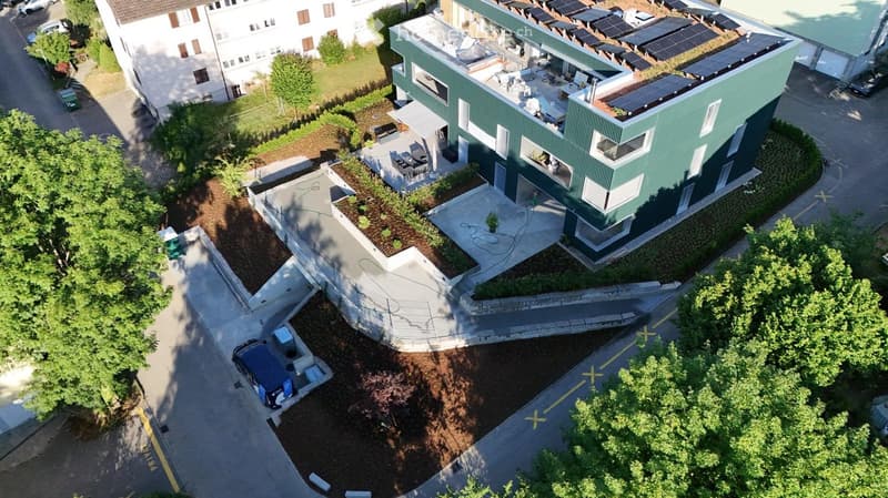 A modern green building with solar panels on the roof, surrounded by landscaped gardens and pathways. The building has multiple levels and large windows, with a raised ground floor and a parking area nearby.