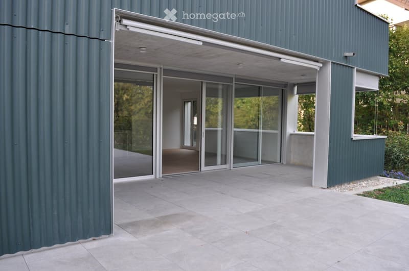 Modern building with large glass sliding doors leading to a covered outdoor area, grey tiled floor, and greenery visible through the glass.