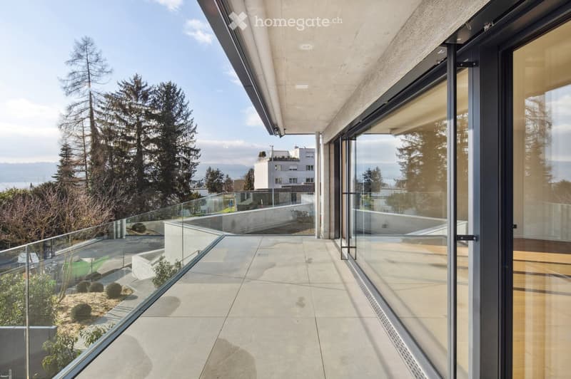 Modern balcony with large glass sliding doors, concrete floor, glass railing, and scenic view of trees and distant mountains.