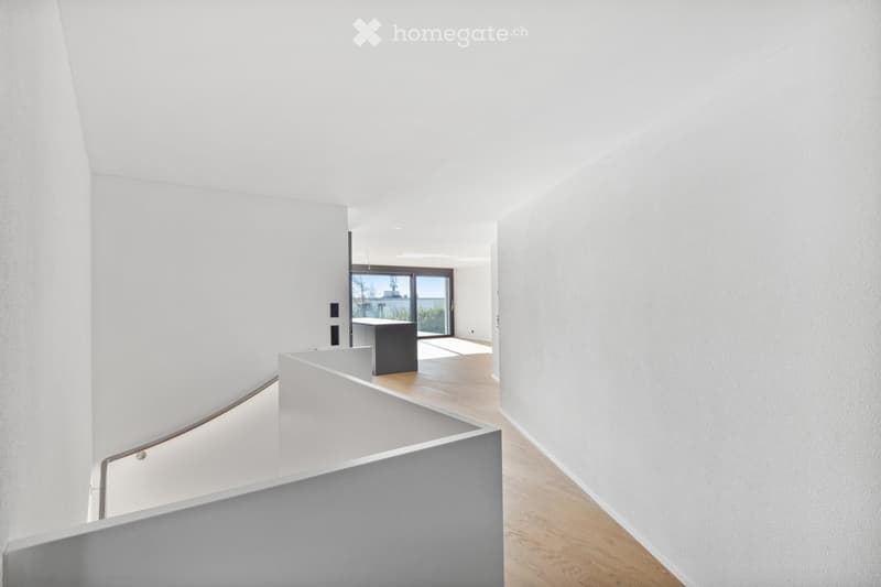 Modern, minimalist hallway with white walls, wooden flooring, and a sleek staircase leading upwards. A small black table is visible in the background near a large glass door opening to an outdoor area.