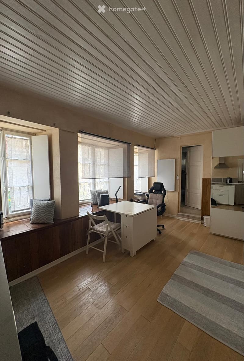 Bright, modern room with wooden flooring, large windows, a desk with chairs, a kitchen area, and a balcony view.