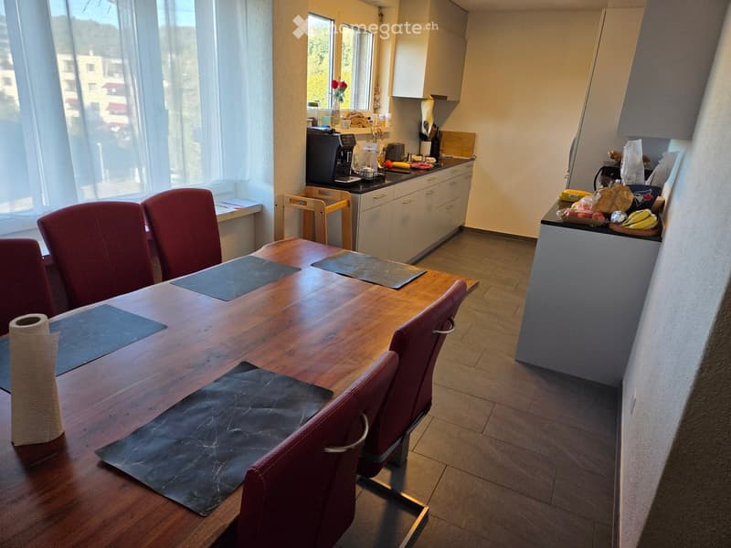 A modern kitchen and dining area with a wooden dining table, red chairs, and various appliances including a coffee maker, fridge, and dishwasher. The area is well-lit with large windows and has a tiled floor.