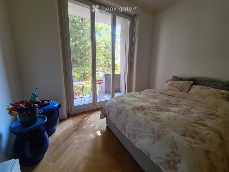Bright bedroom with wooden flooring, a large bed with floral bedding, a blue nightstand with a vase of flowers, and a sliding glass door leading to a balcony with outdoor furniture.