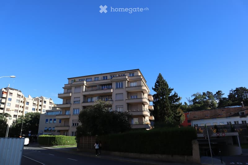 Multi-story residential building with balconies and large windows, surrounded by trees and greenery.