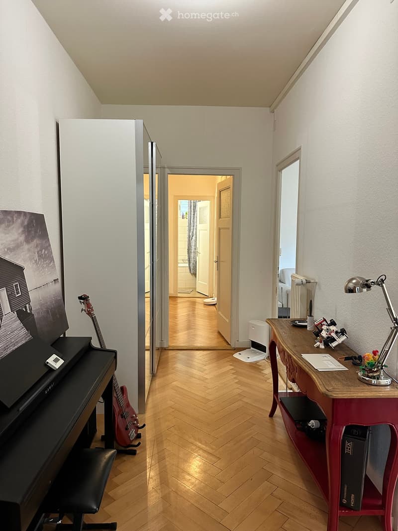 A hallway with herringbone wooden flooring, featuring a piano with a stool, a guitar, a wooden table with a lamp, and a mirror on the wall.