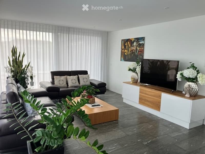 Modern living room with large windows, dark grey sectional sofa, wooden coffee table, TV stand, and decorative vases with flowers.