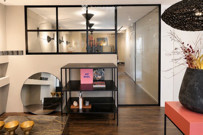 Modern hallway with wooden flooring, large glass windows, a black metal shelf with books and decorative items, a pink table with a large black vase, and several golden bowls on the floor.